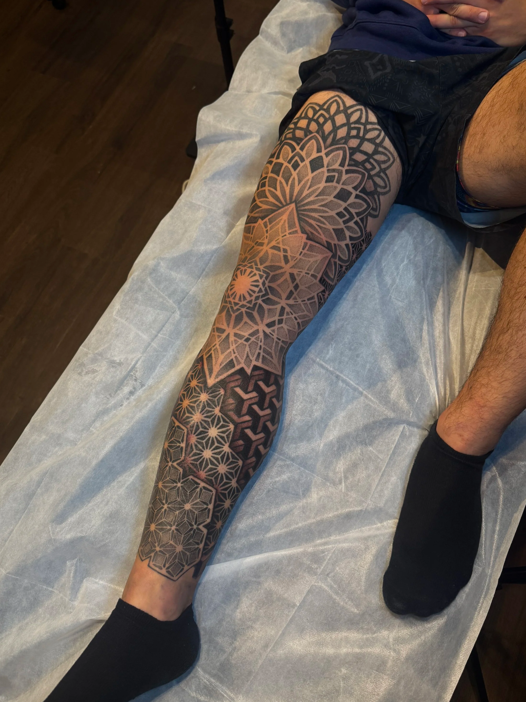 A person's arm with a large geometric tattoo, resting on a padded surface, wearing black shorts and a dark-colored shirt.