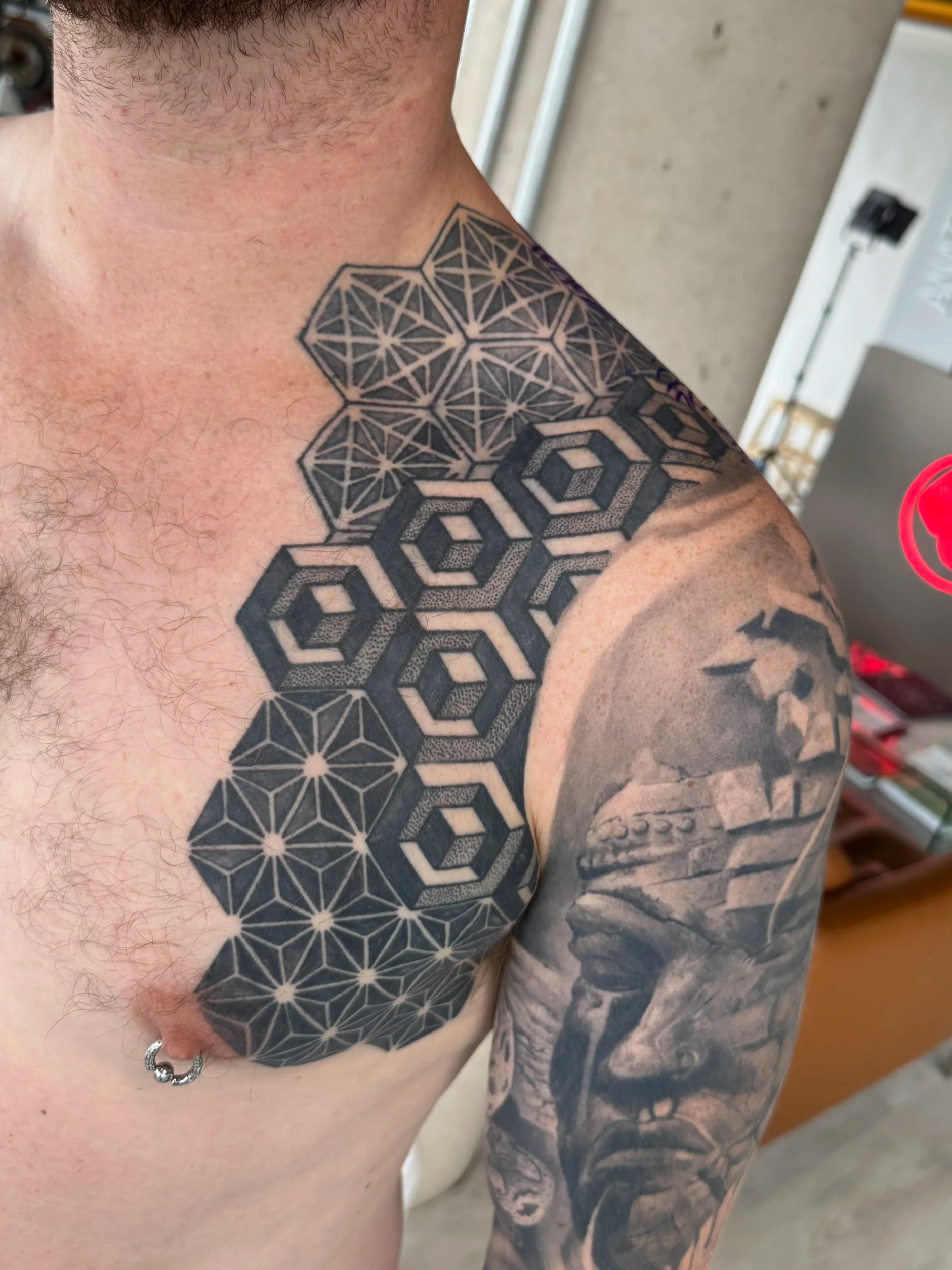 Tattoo of geometric patterns on a person's shoulder and upper sleeve, primarily black ink, with some shading and line work, including interconnected hexagons and star-like shapes.