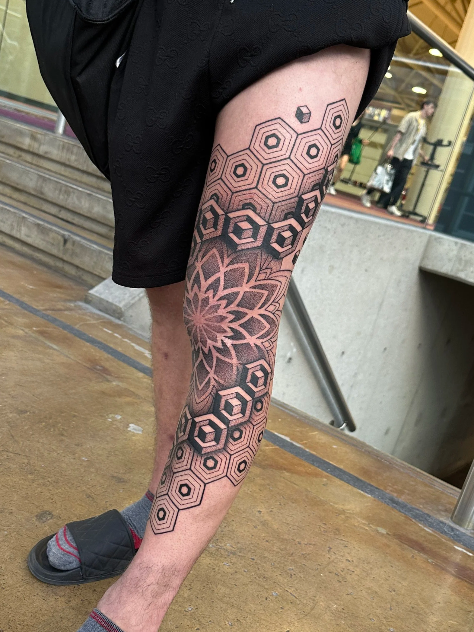 A person's leg with black geometric tattoo designs, wearing dark shoes and striped socks, standing on a wooden and concrete floor inside a building.