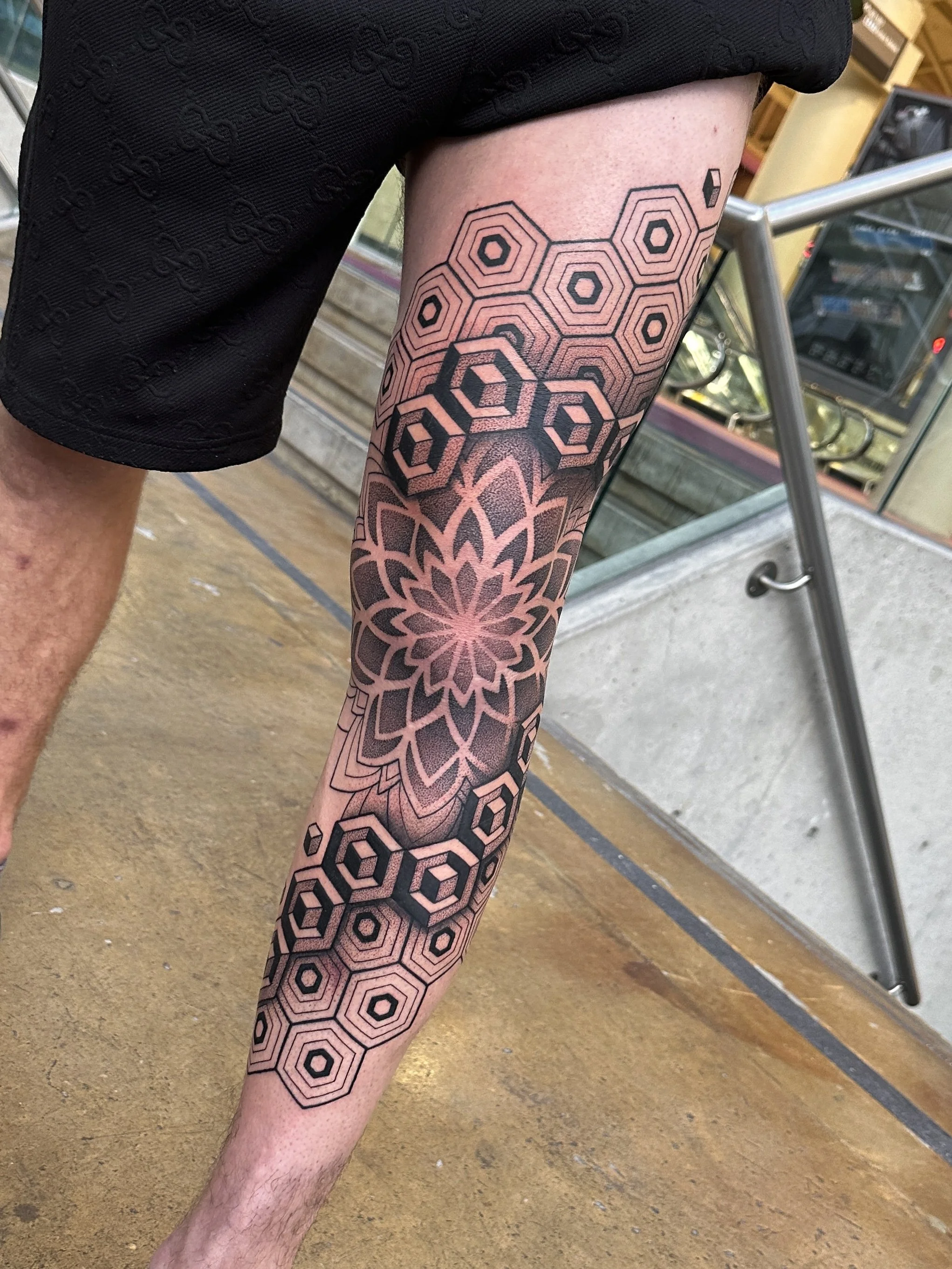 A person's leg with a geometric tattoo design featuring hexagons and a floral pattern, while wearing black shorts, in an indoor setting with a metal railing and store shelves in the background.