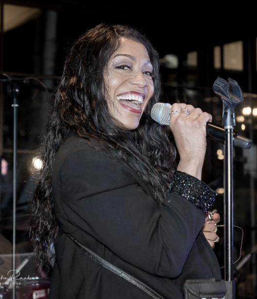A woman performing with a microphone in her hand, smiling and looking to the side, wearing a black blazer with glittery sleeve cuffs, and long dark hair.