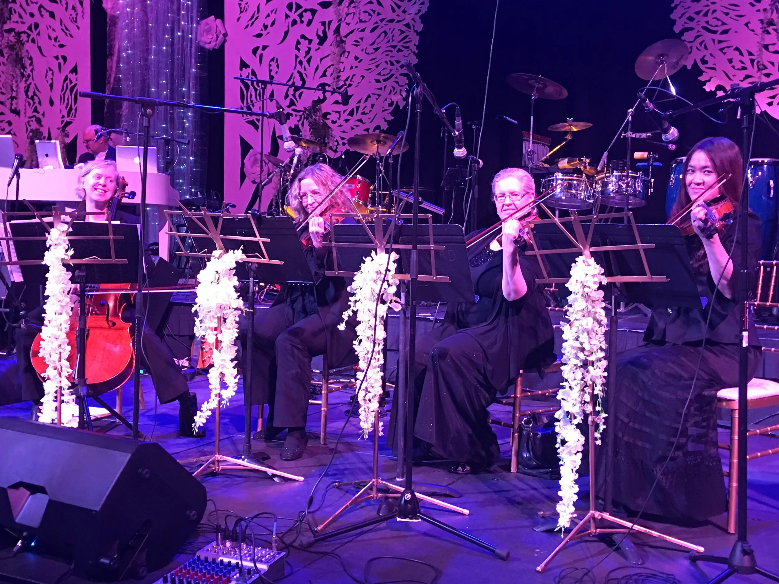 A group of musicians performing on stage with various instruments, including violins, drums, and a keyboard. The stage is decorated with floral and tree motif designs in the background, illuminated with purple and pink lighting.