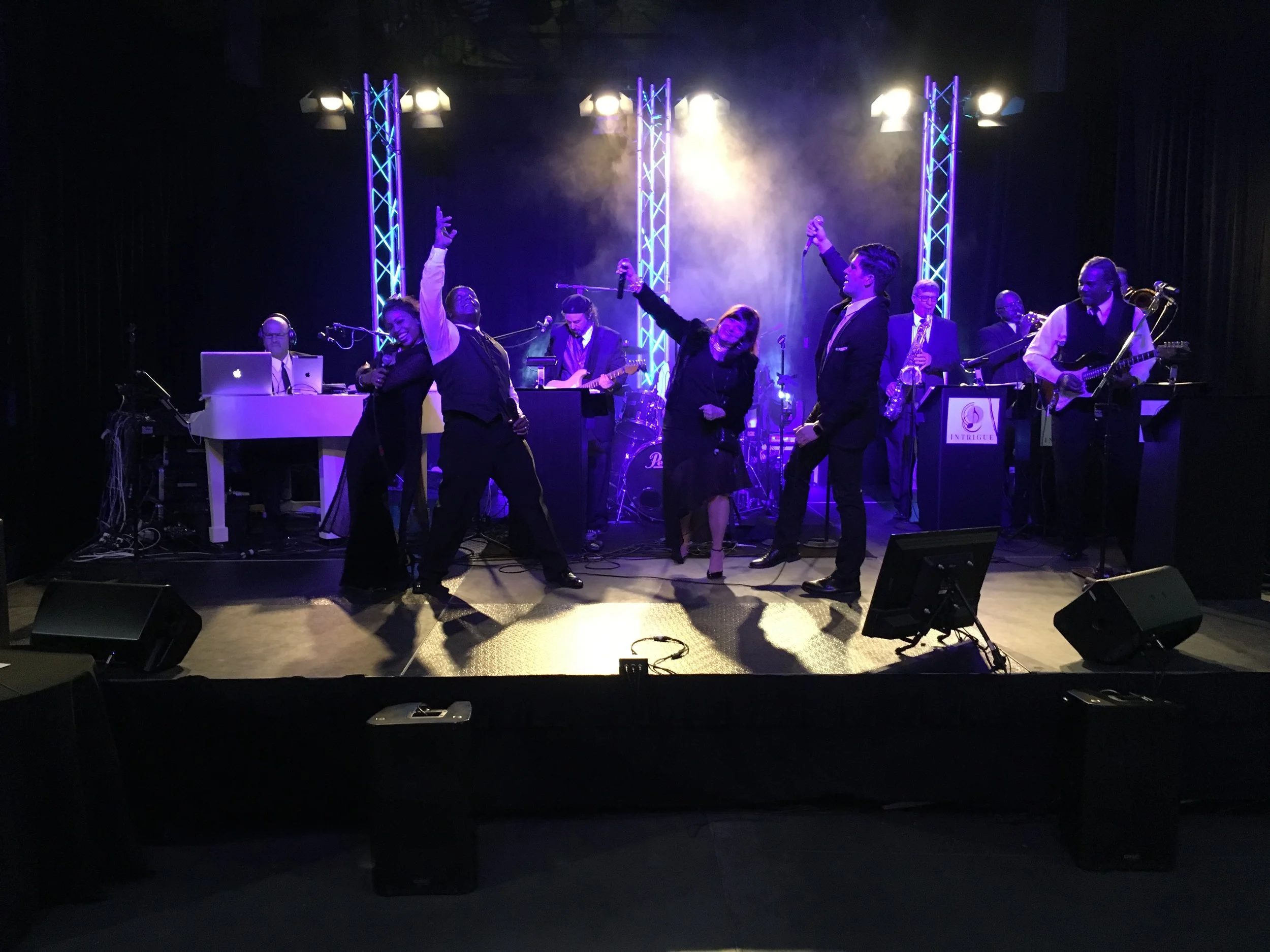 Musicians performing on stage with blue and purple lighting, including a pianist, singers, guitarists, and a saxophonist, with a black backdrop and sound equipment in front.