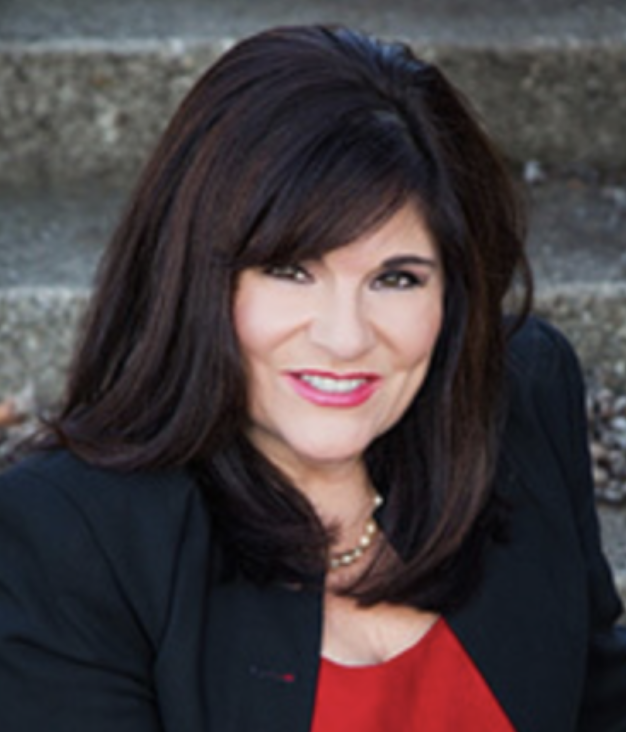 A woman with shoulder-length dark brown hair, wearing a black blazer and a red top, smiling outdoors with steps in the background.