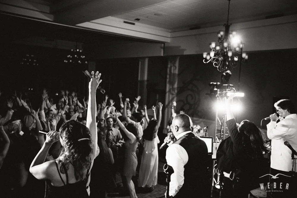A lively crowd dancing and singing at a party or concert, with multiple performers on stage holding microphones and singing, in a black-and-white photo.