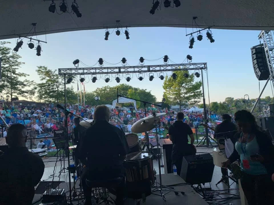 View of a stage with musical instruments and performers facing an outdoor audience during daytime, with trees and blue sky in the background.