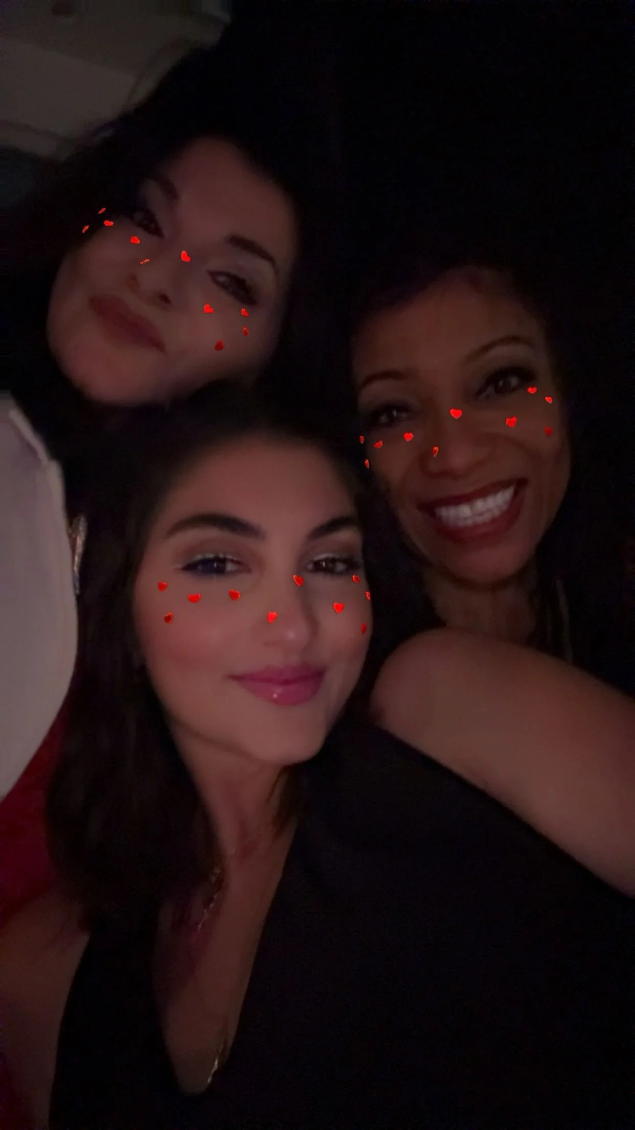Three women taking a selfie, each with small red heart icons on their faces, in a dark setting.