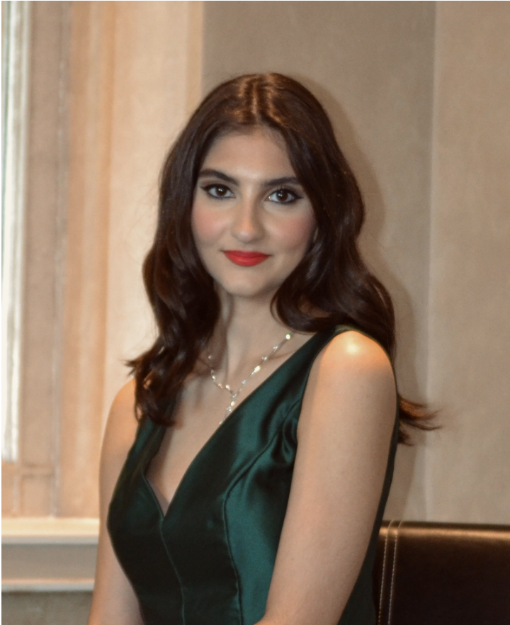 A woman with dark brown hair wearing a dark green satin sleeveless dress and a necklace, posing indoors with a neutral background and soft lighting.