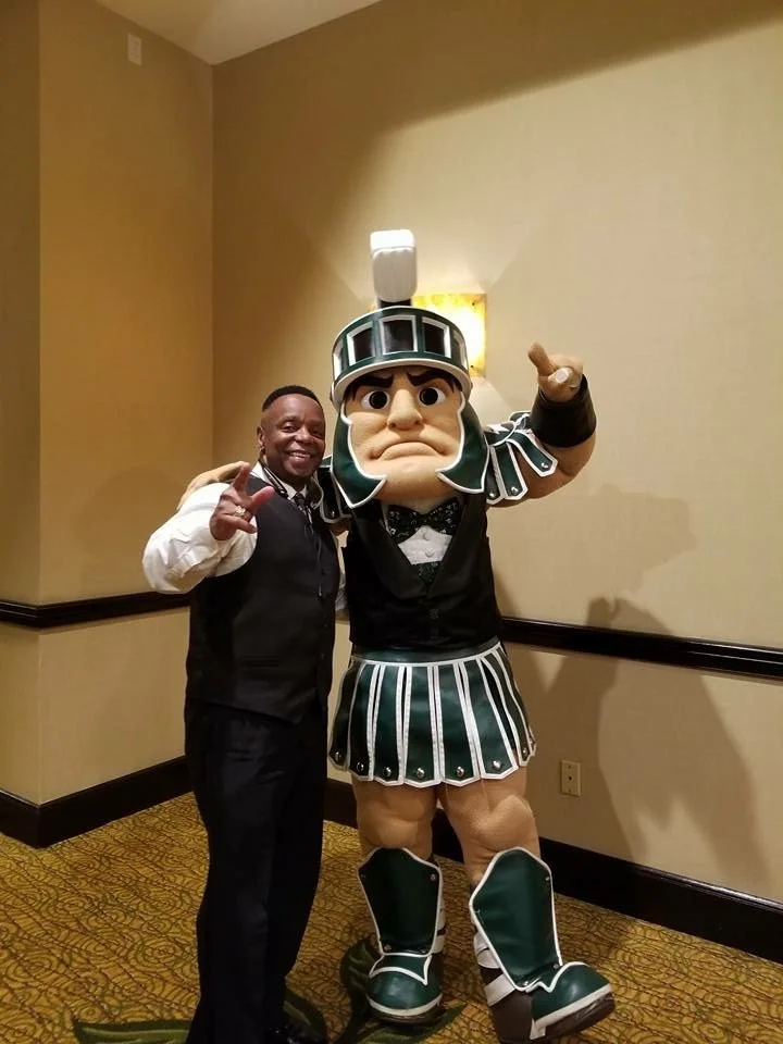 A man in a formal vest and shirt standing next to a person in a large mascot costume resembling a football player in a green and white uniform. The mascot has a serious expression and is pointing forward.