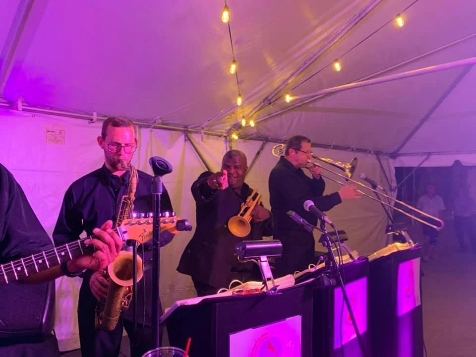 Jazz band performing under a tent with string lights, featuring musicians playing saxophones, trumpet, and guitar.