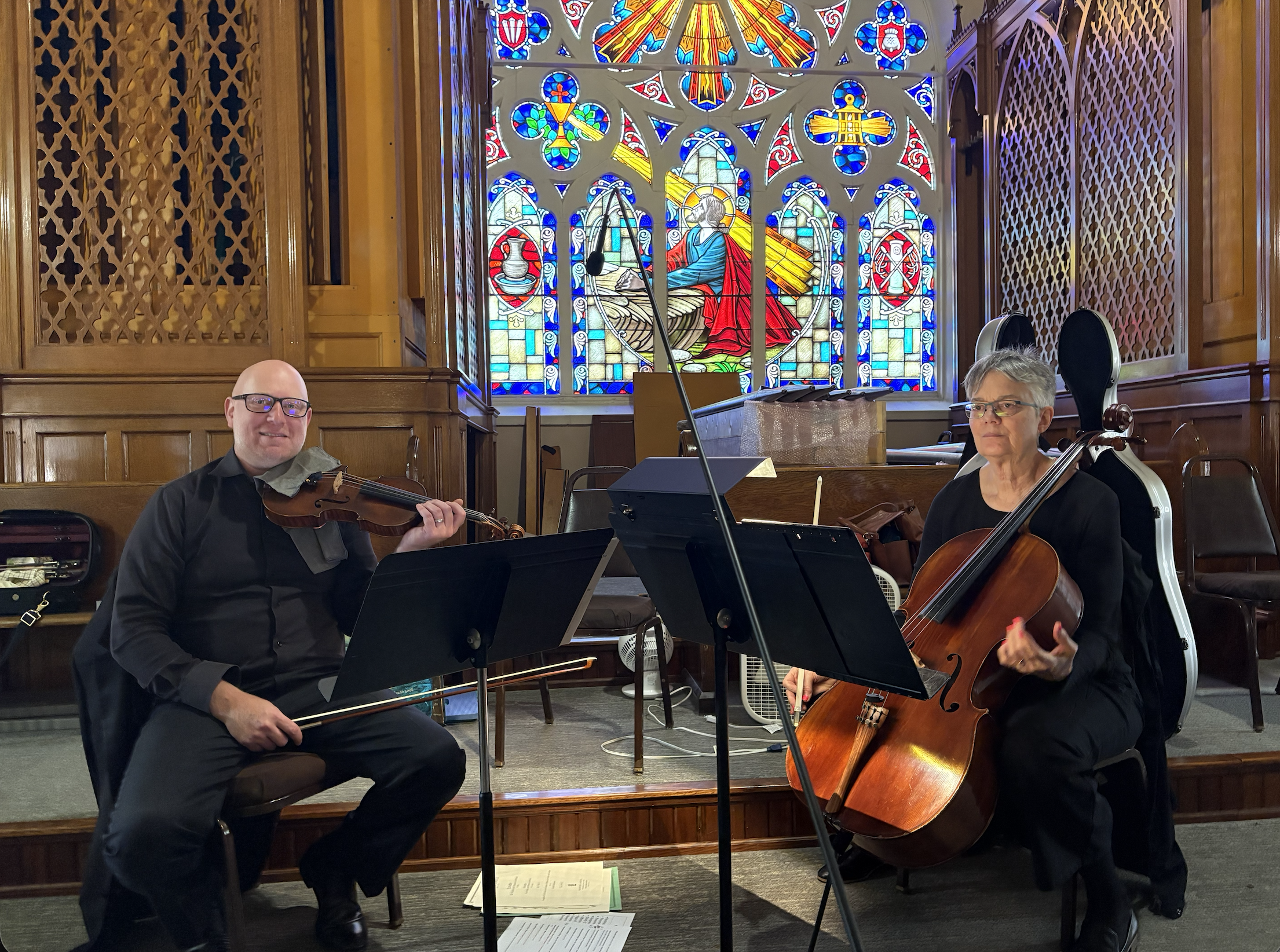 Two musicians are playing instruments in a church with stained glass windows. The man on the left is playing a violin, wearing glasses and a black shirt. The woman on the right is playing a cello, wearing glasses and a black top.