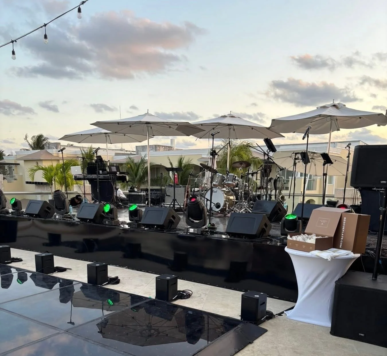 An outdoor stage setup for a concert with musical instruments, including drums, keyboards, and microphones, under large umbrellas. There are stage lights, speakers, and some boxes on a table to the side, with a scenic background of buildings, trees, and a cloudy sky.