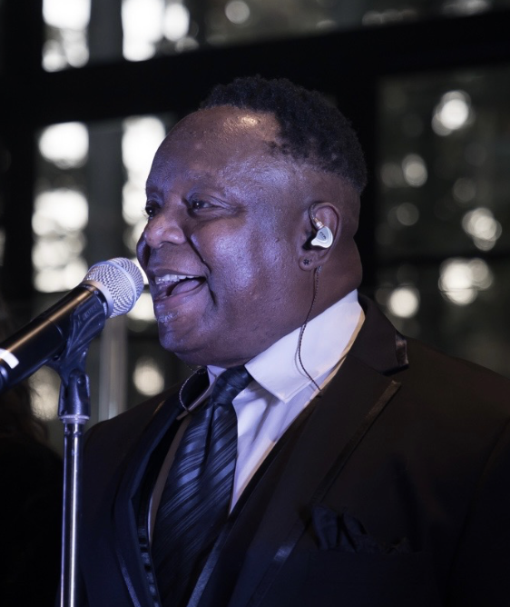 A man in a black suit and tie singing into a microphone at a live event.