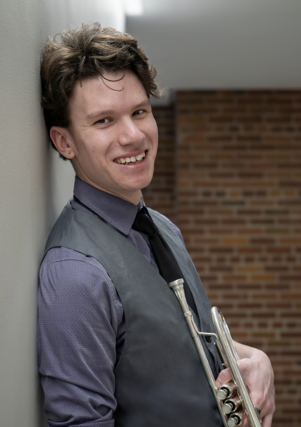 Young man with curly hair leaning against a wall, holding a trumpet and smiling.