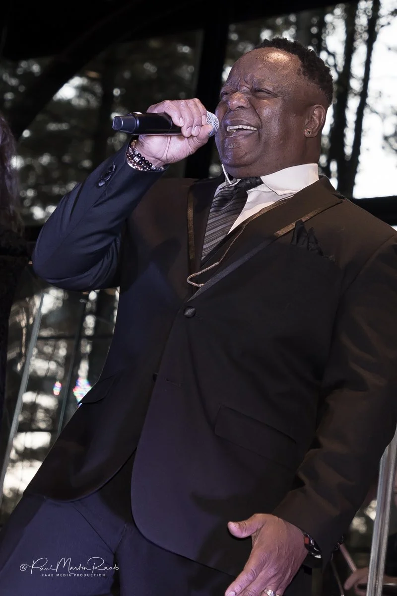 A man in a suit singing into a microphone during a performance or event, with trees visible in the background.