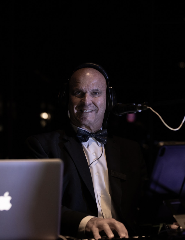 A man dressed in a tuxedo and bow tie, wearing headphones, is smiling while performing music at a microphone with laptops nearby in a dark setting.