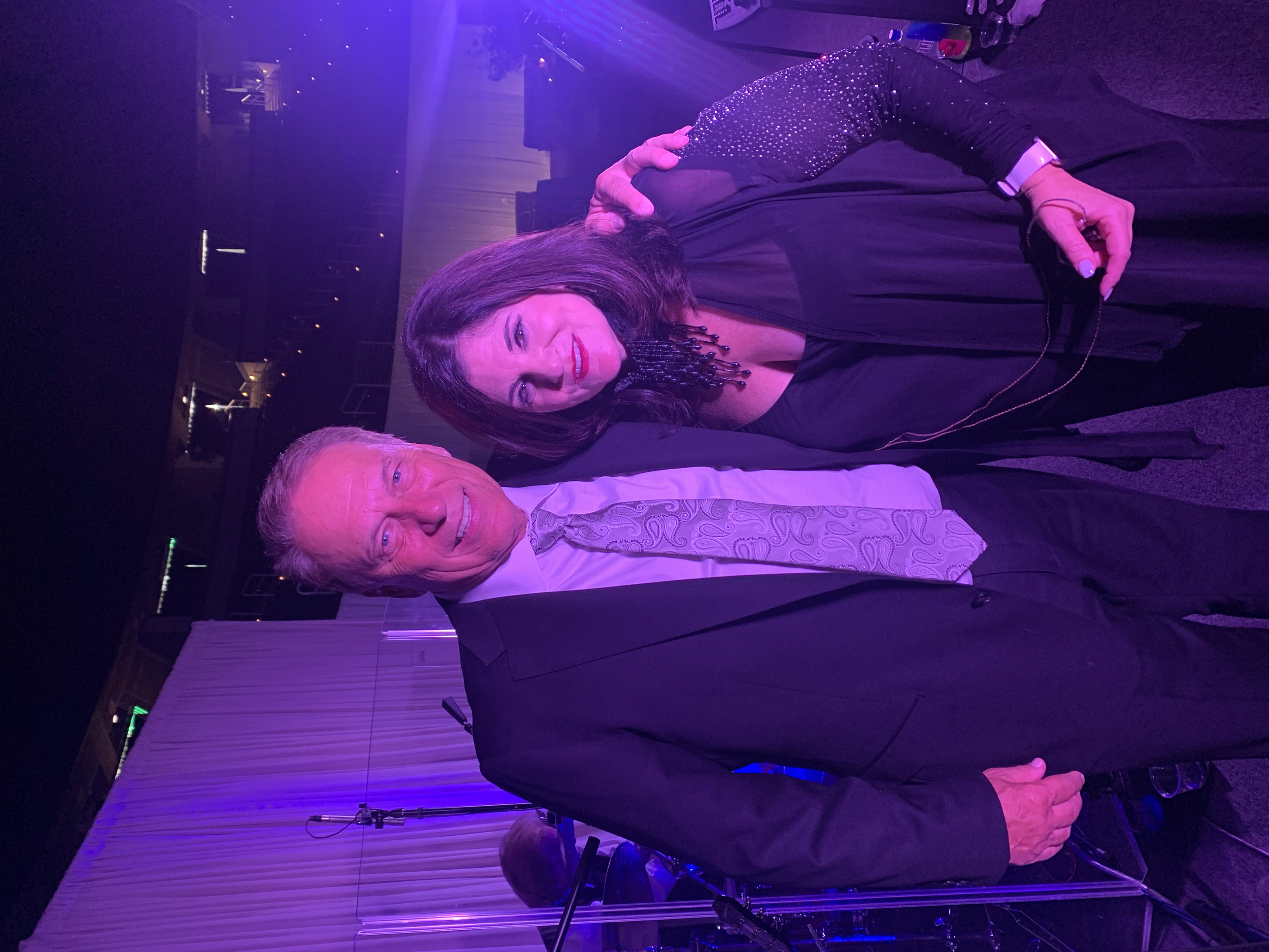 A woman with dark hair and a black dress, adorned with sparkles, standing next to a man with gray hair in a white shirt and patterned tie, at an indoor event with purple lighting and a stage in the background.