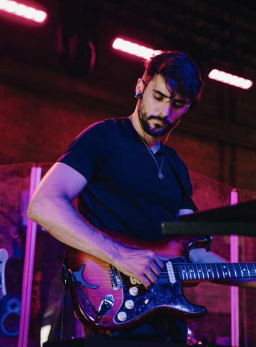 A man with dark hair and beard playing an electric guitar on stage with pink and purple lighting.
