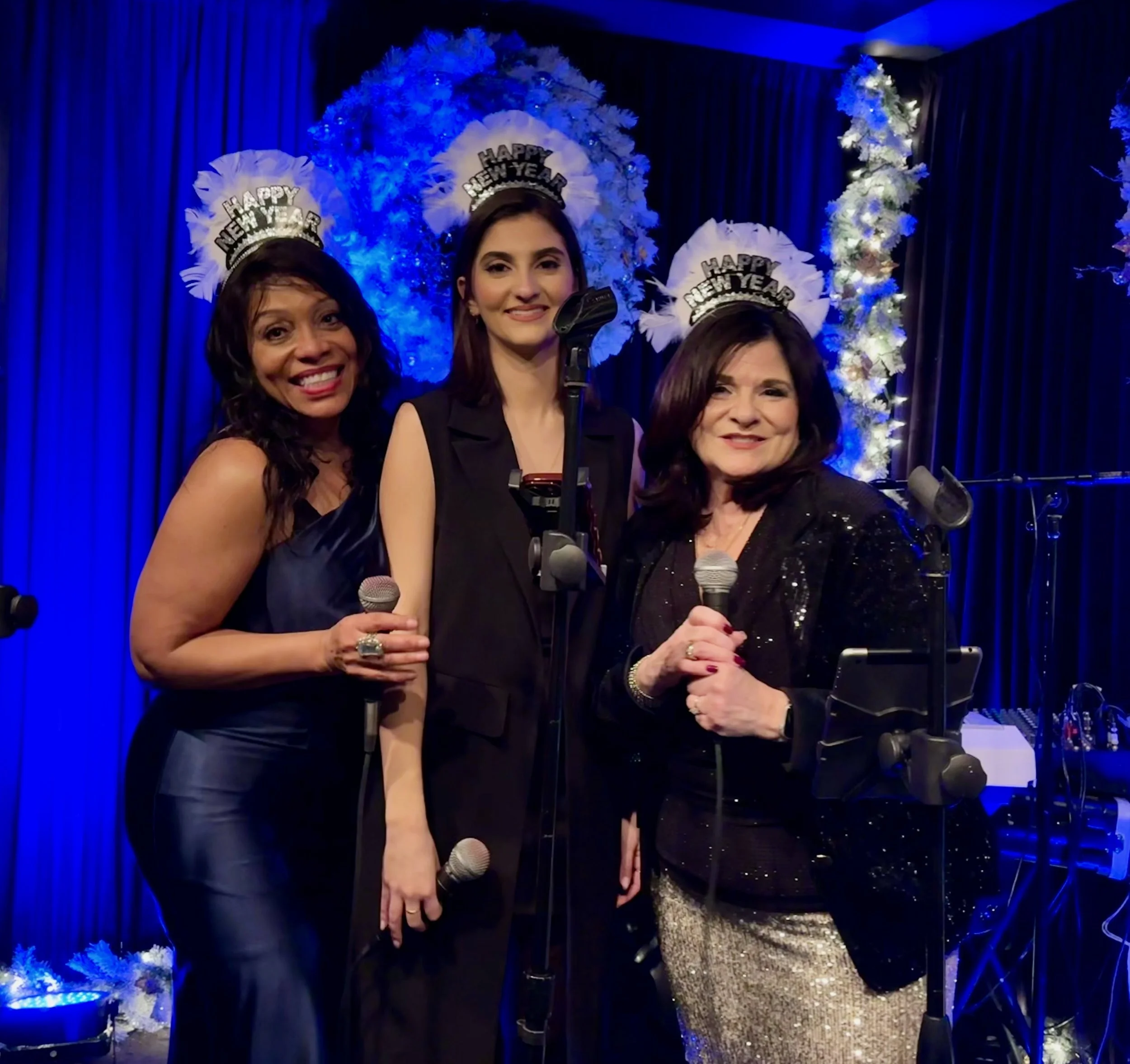 Three women celebrating New Year's Eve, wearing paper hats with 'Happy New Year' written on them, holding microphones, standing in front of a festive blue and white floral backdrop with string lights.