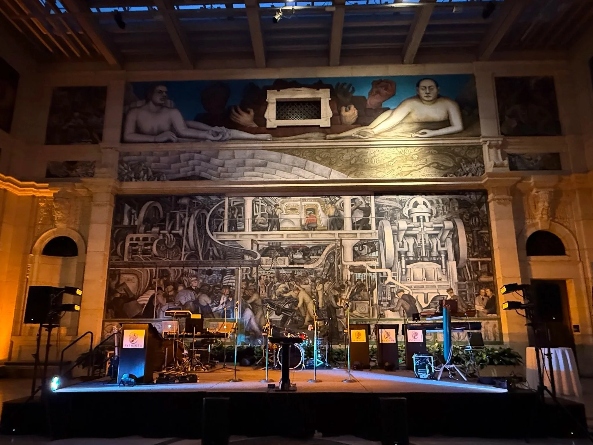 Interior view of a historic building with mural paintings on the upper wall, depicting human figures and machinery, and a stage with musical instruments and equipment in the foreground.