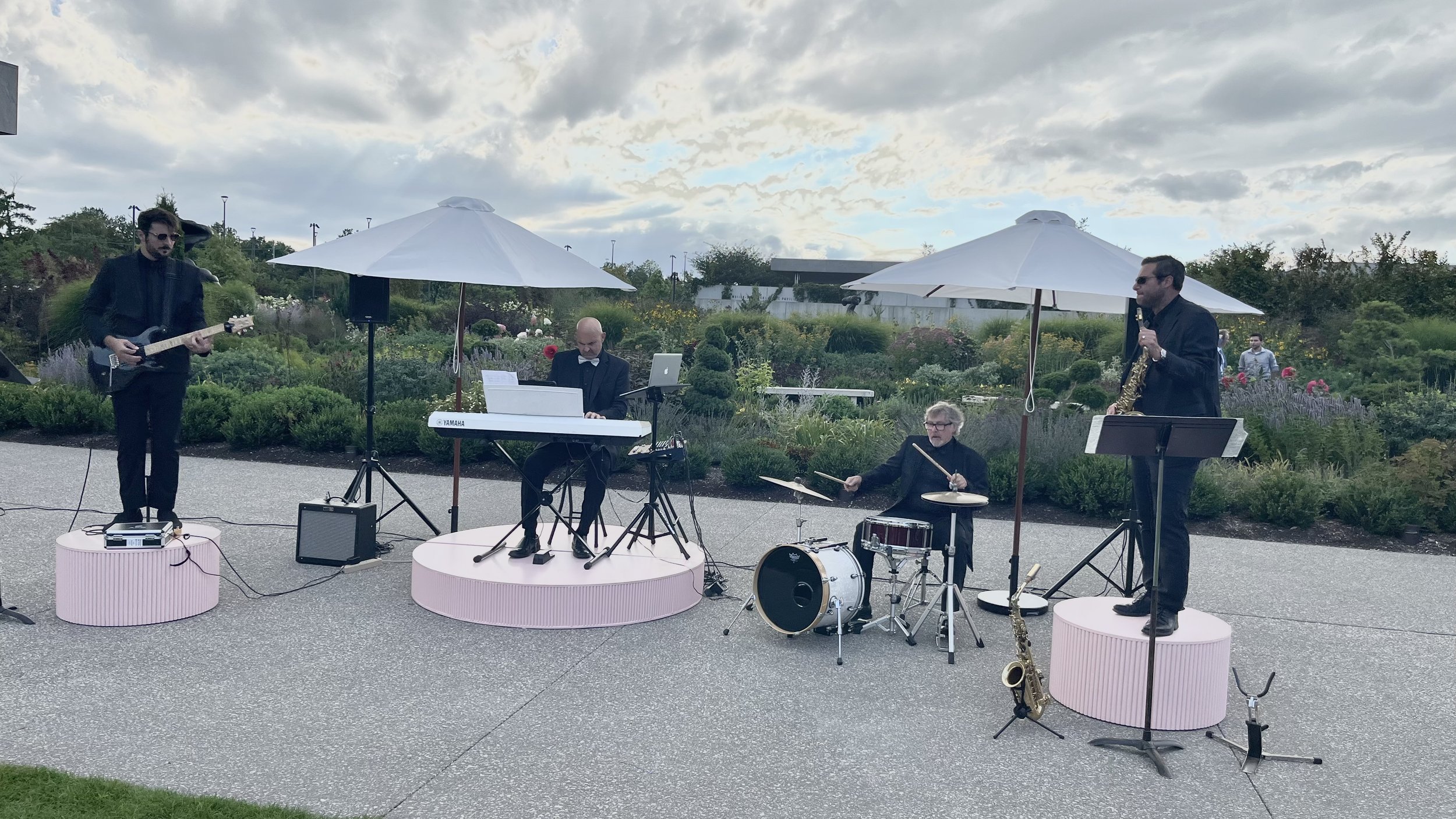 A band of four musicians performing outdoors on a paved area with garden in the background. One playing guitar, one on keyboard, one on drums, and one on saxophone. They are dressed in black, with umbrellas providing shade.