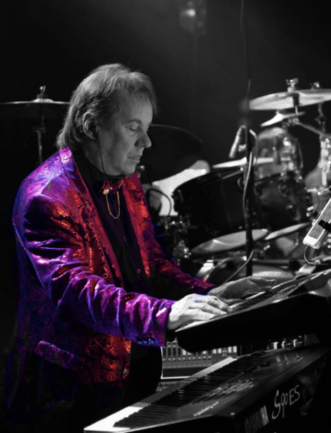 A man playing a keyboard on stage, wearing a red and purple patterned jacket.
