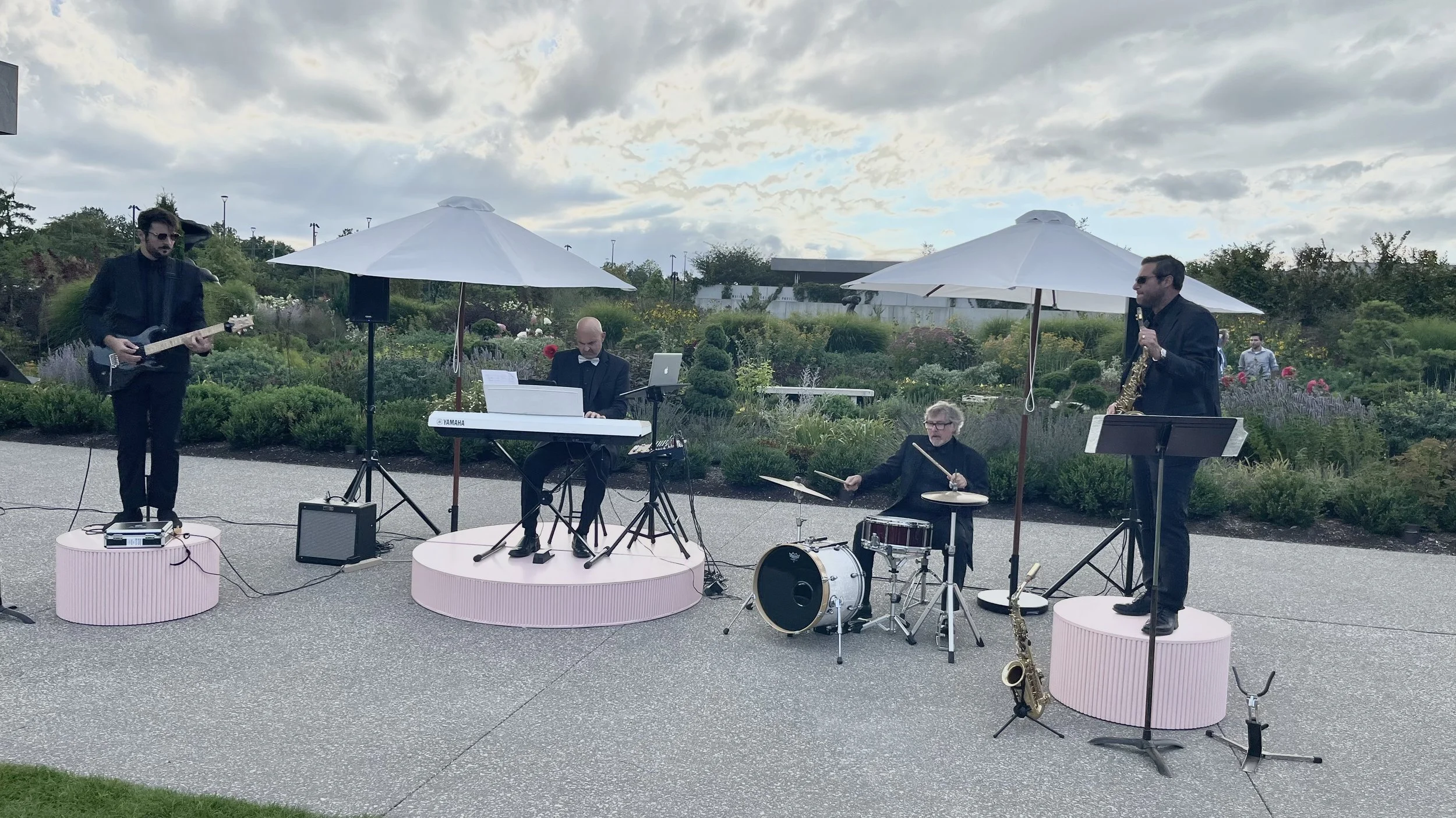 A band performing outdoors on a paved area surrounded by greenery and flower bushes, with cloudy skies overhead. The band includes a guitarist, a pianist, a drummer, and a saxophonist, all dressed in formal black attire. Small pink platforms elevate 