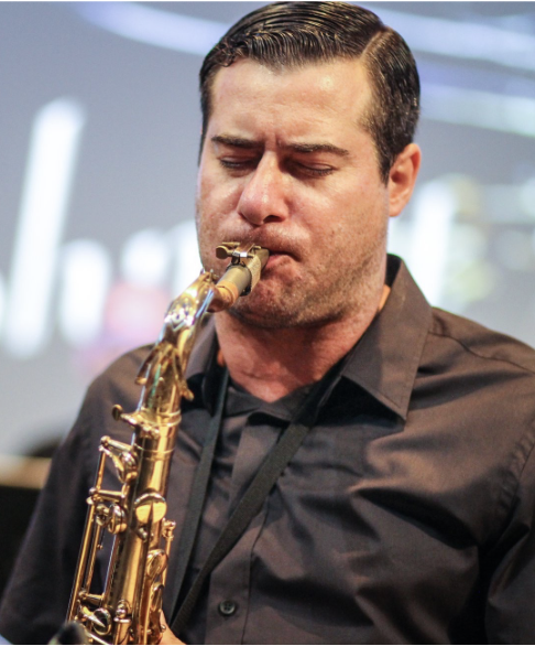 Man playing a gold saxophone, with eyes closed, in an indoor setting with blurred background.