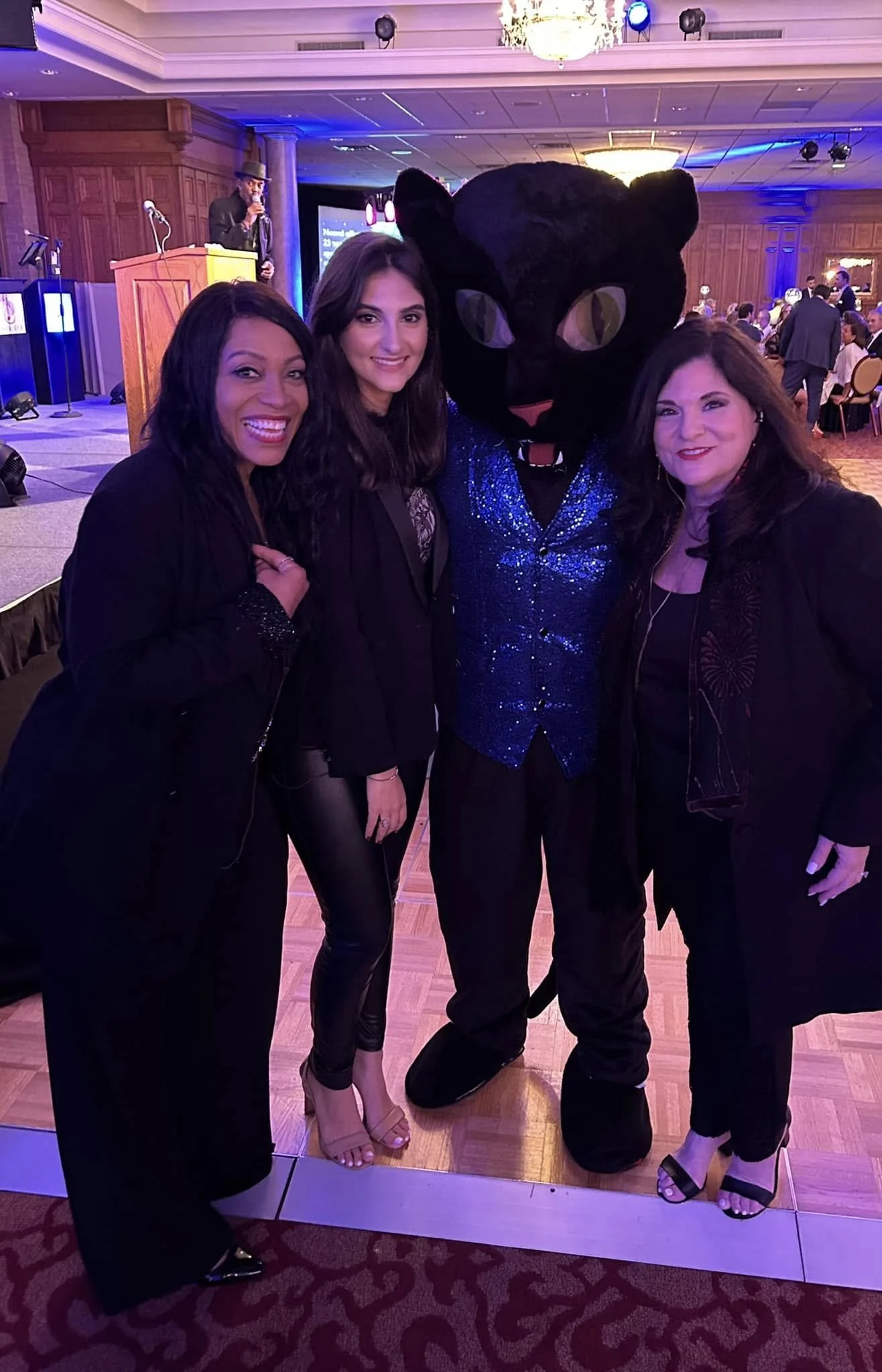 Women and a person in a panther costume posing at an event with a stage, a speaker at a podium, and a group of people seated in the background, all inside a large decorated ballroom.