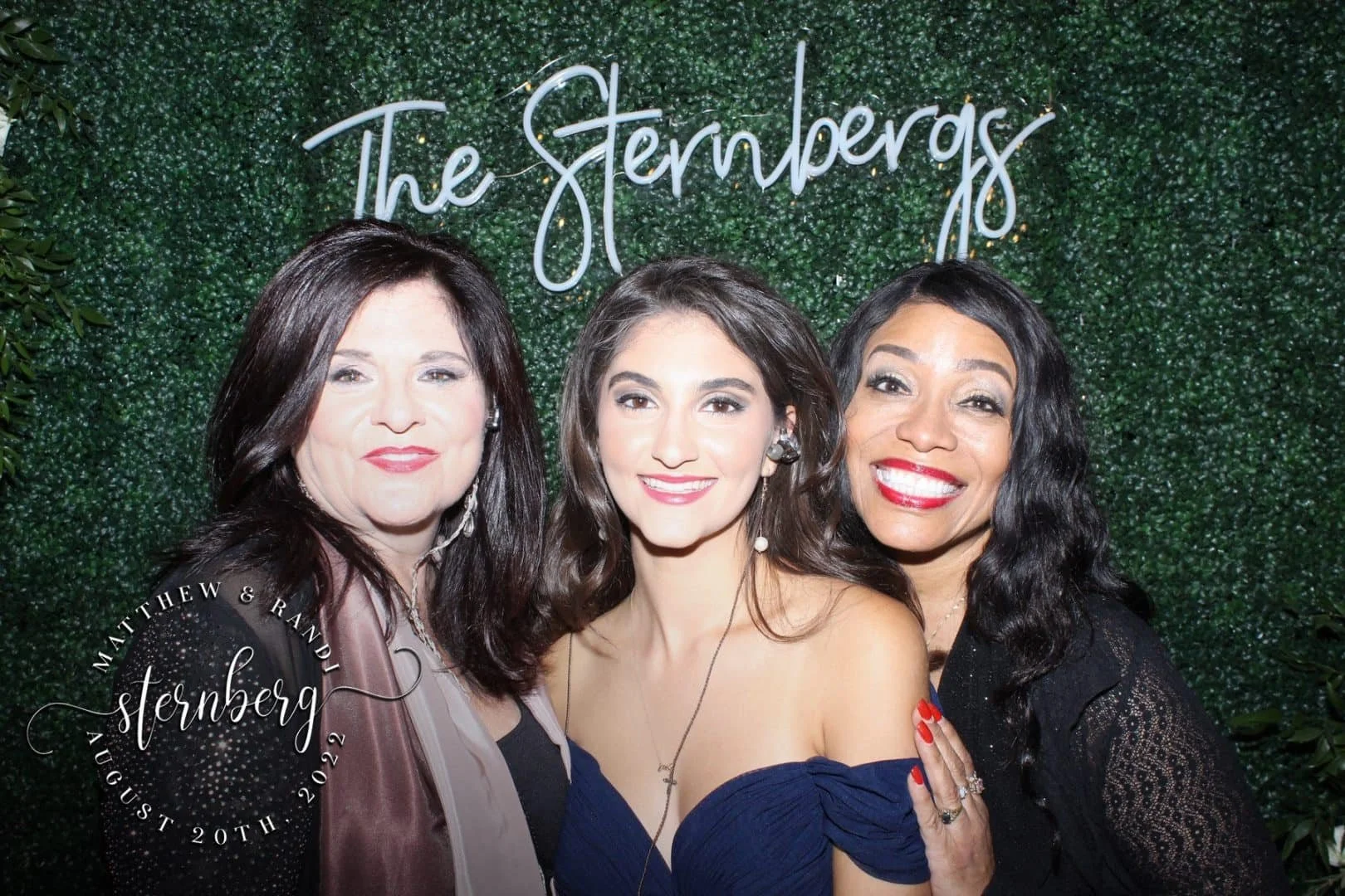 Three women smiling at a photoshoot with a green leafy background and a neon sign that reads 'The Sternbergs.' One woman has dark brown hair, the second has medium brown hair, and the third has black hair with waves. They are dressed in elegant clothing and jewelry.