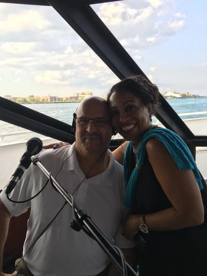 Two people, a man with glasses wearing a white shirt and a woman with dark hair and a blue dress, smiling together on a boat with water and a distant city skyline in the background.