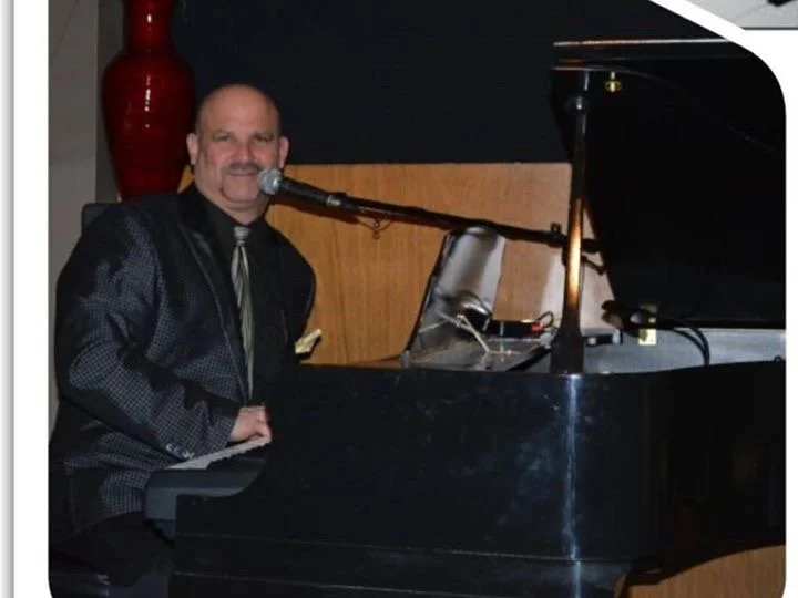 A man in a dark suit playing a grand piano and singing into a microphone in a dimly lit room.