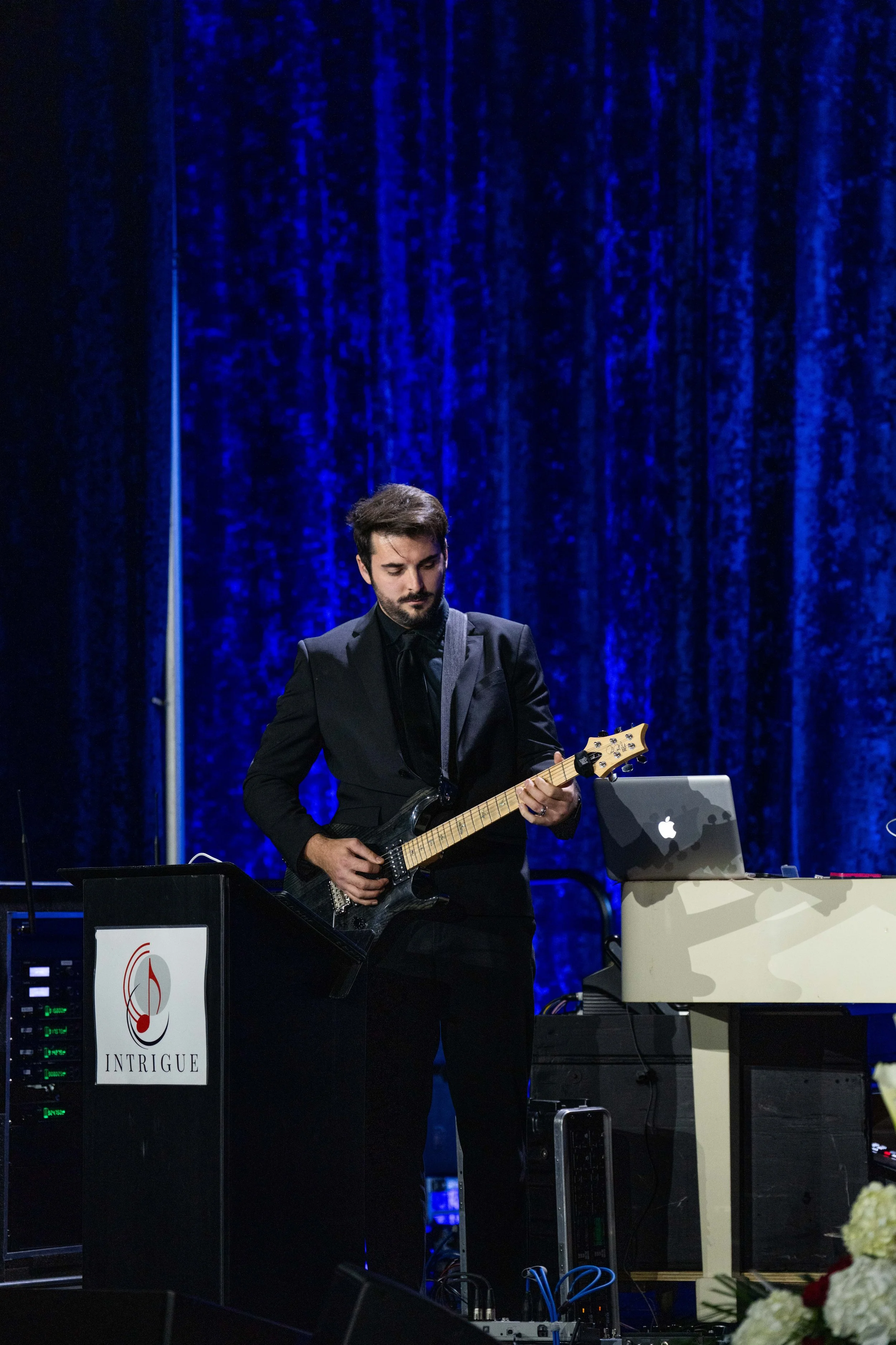 A man wearing a black suit playing an electric guitar on a stage with a deep blue curtain backdrop, with a white keyboard and a silver MacBook computer nearby, and flowers in the foreground.