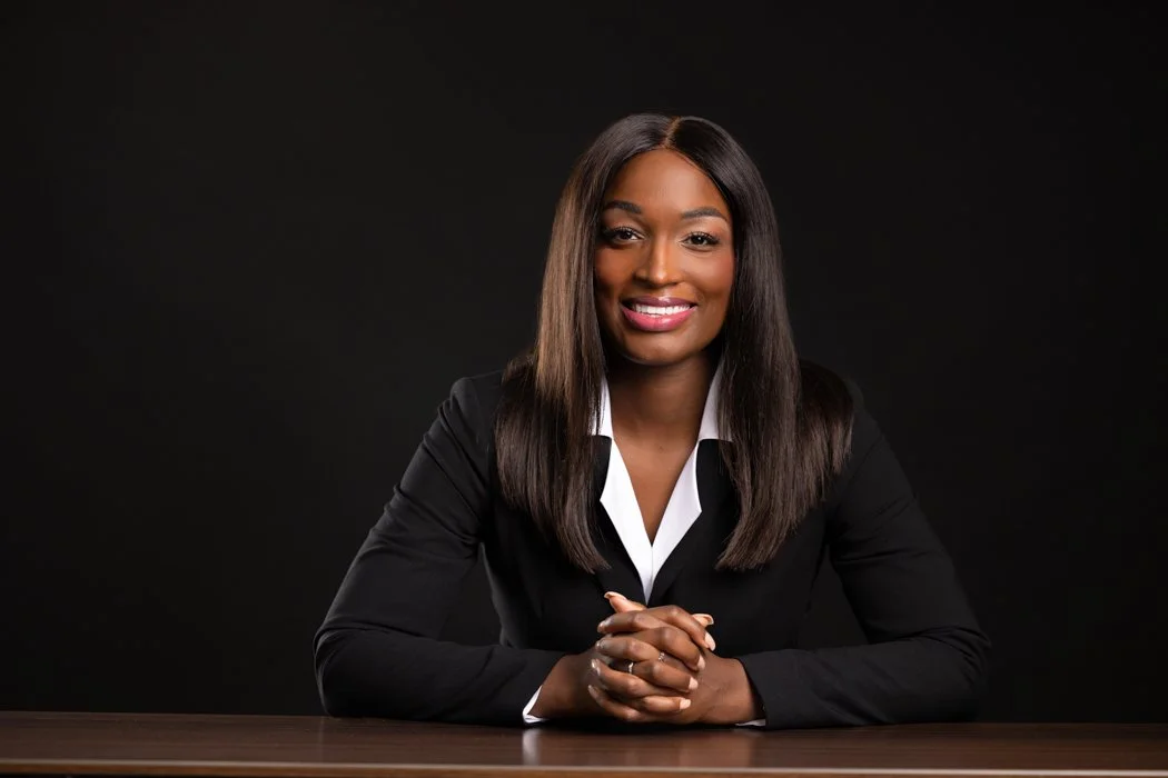 Professional woman with long dark hair, wearing a black blazer and white shirt, sitting at a table against a dark background.
