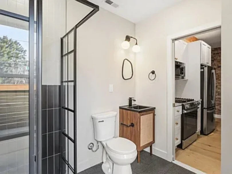 Bathroom with shower, toilet, small wooden vanity with black sink, wall mirror, and light fixture; kitchen with stove, refrigerator, and white cabinets visible through doorway.