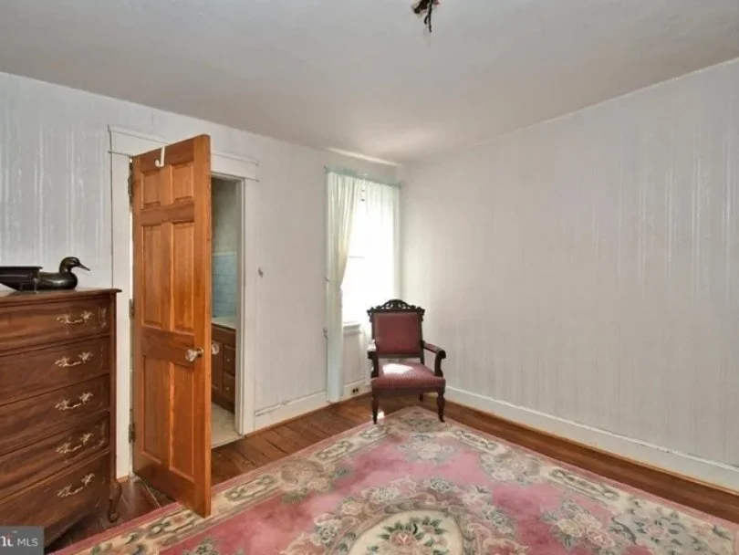 A simple bedroom with white paneled walls, wooden floor, and a pink floral area rug. There is a wooden dresser with a duck-shaped decorative item on top and an armchair with dark wood frame and pink upholstery near a window with light curtains. The d