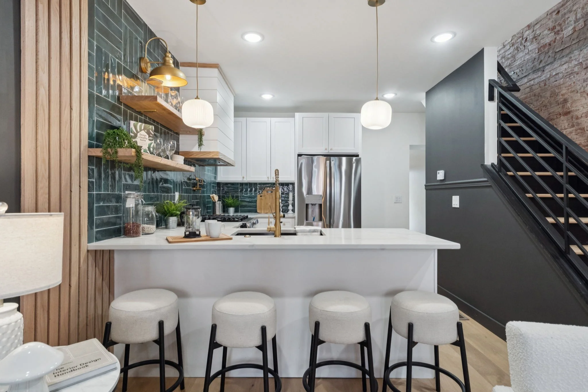 Interior view of a modern kitchen with white cabinets, stainless steel refrigerator, green tiled backsplash, and wooden shelves with glassware and plants. Pendant lights hanging above a white island with four bar stools in front. Black staircase with