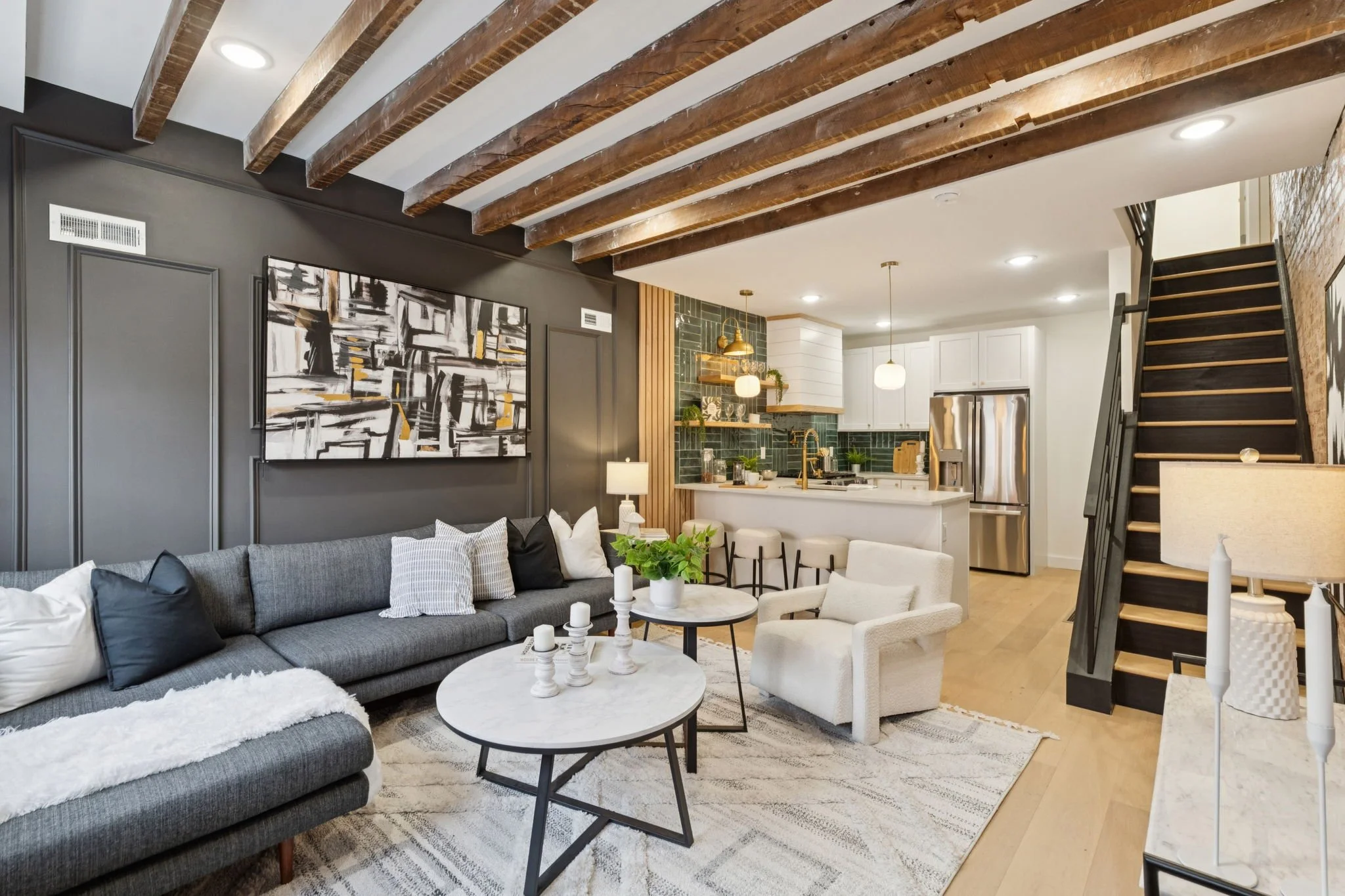 Open concept living room and kitchen with exposed wooden ceiling beams, gray and white furniture, modern artwork, and hardwood floors.