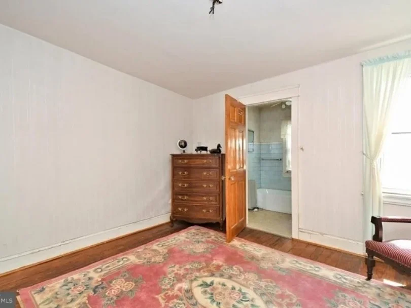 Empty bedroom with a wooden dresser, a door leading to a bathroom with light blue tiles, a window with light-colored curtains, and a patterned pink and green rug on wooden floors.