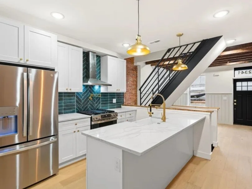 Modern kitchen with white cabinets, teal backsplash, stainless steel appliances, and a white marble island with gold fixtures.