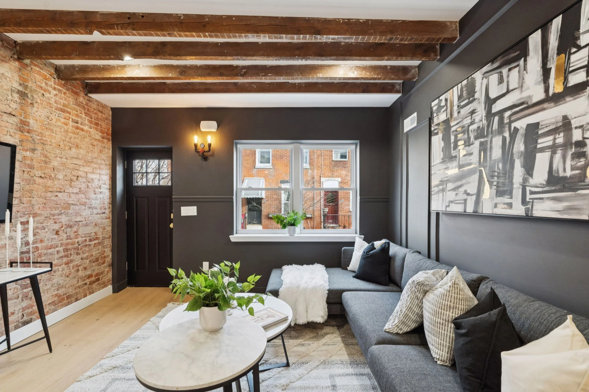A living room with exposed brick and dark painted walls, wooden ceiling beams, a window with a potted plant on the windowsill, a gray sectional sofa with patterned and solid pillows, a large abstract black and white painting, a white round coffee tab