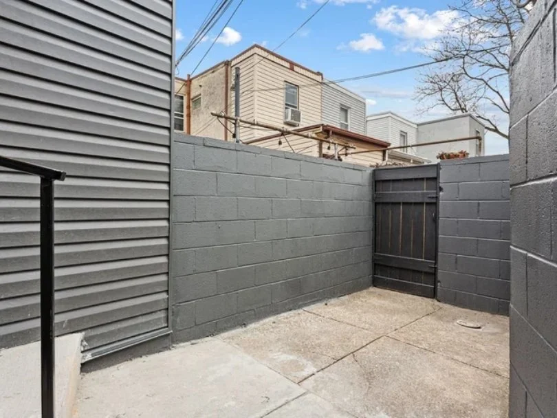 Small backyard enclosed by gray brick walls, with a black wooden gate and neighboring buildings visible in the background under a partly cloudy sky.