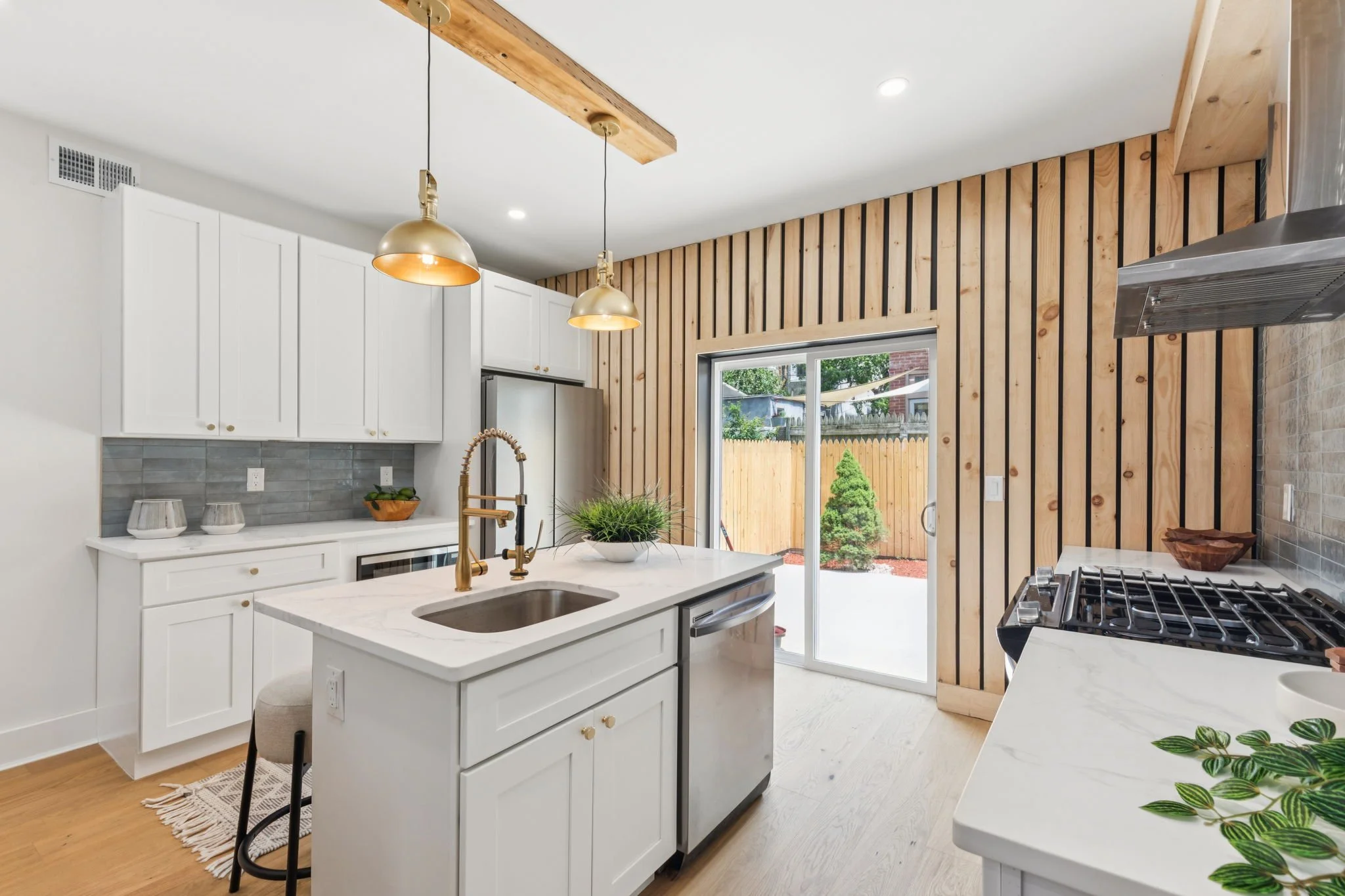 Modern kitchen with white cabinets, marble island, wood panel accent wall, stainless steel appliances, pendant lights, sliding glass door showing backyard with trees and fence.