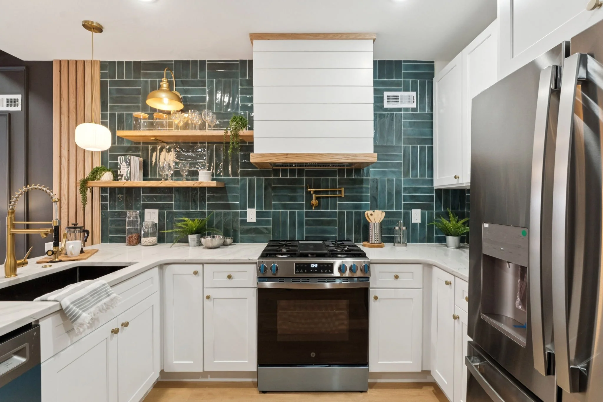 A modern kitchen with white cabinets, a stainless steel stove, green tile backsplash, wooden shelves, gold accents, and a stainless steel refrigerator.