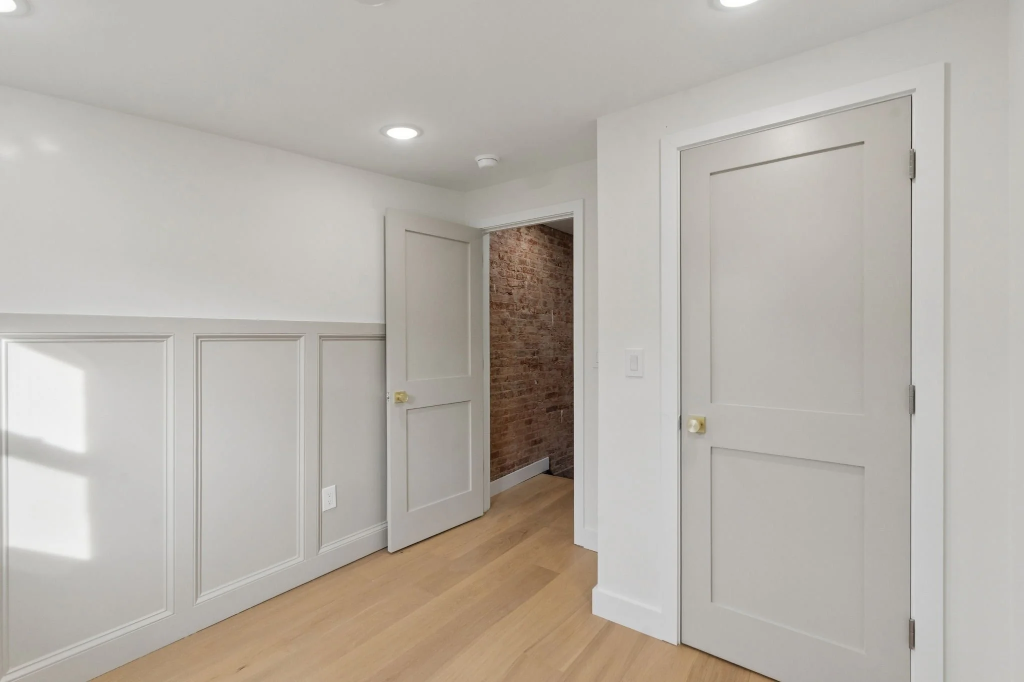 Interior view of a room with white walls featuring decorative paneling, a closed white door, a partially open door revealing an exposed brick wall, wood flooring, and ceiling lights.