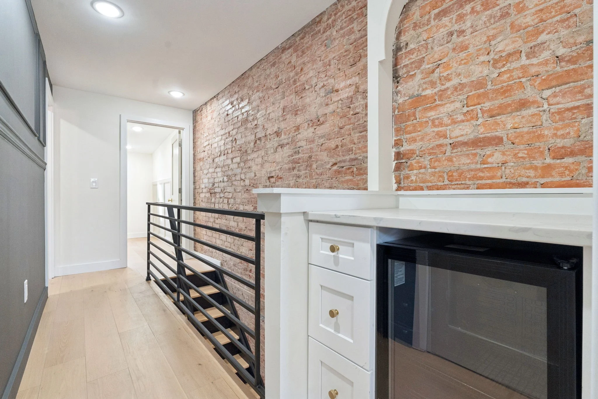 Interior view of an apartment hallway with exposed brick wall, white trim, and a white fireplace mantel. There is a black metal railing in the foreground, and a door at the end of the hallway.
