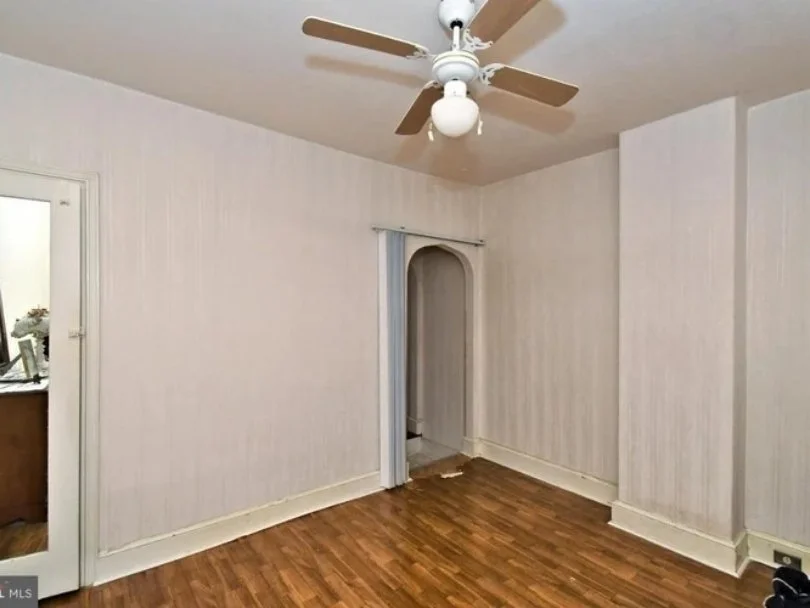 Empty room with beige walls, a ceiling fan with a light, a partially open archway with vertical blinds, and hardwood flooring.