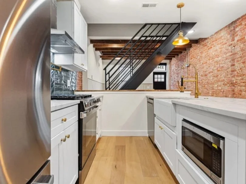 Modern kitchen with white cabinets, brass fixtures, stainless steel appliances, exposed brick wall, wooden flooring, and a black staircase.
