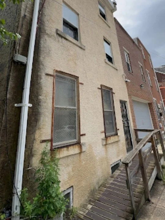 Side of a weathered yellow building with three windows, some with rusted metal braces, a downspout, a small green plant, and a wooden ramp leading to a door.