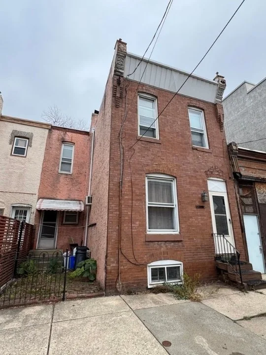 A three-story brick row house with one large window on the first floor, two smaller windows on the second floor, and one small basement window. There are stairs leading up to a door on the right side, and a small, fenced yard area on the left with so